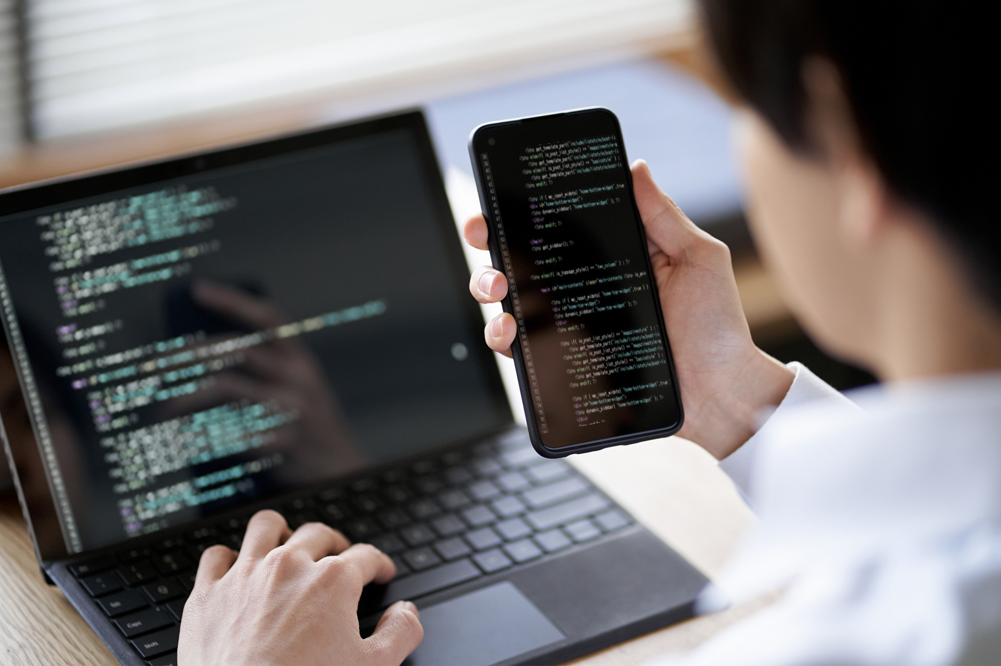 Person at computer looking at code and holding cell phone for mobile testing